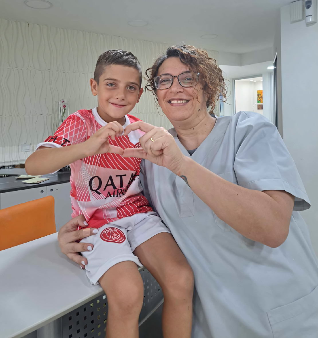 Dr. Yiota and a child in the dental center, smiling as they form a heart shape with their hands while the child sits on a counter.